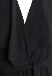 Black layered blouse with a textured finish, featuring a front tie detail and v-neck design, emphasizing a smooth, elegant fabric.