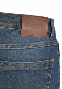 Poche arrière d'un jean en denim bleu avec des coutures orange et un patch en cuir marron portant la marque "HECHTER" près de la taille.