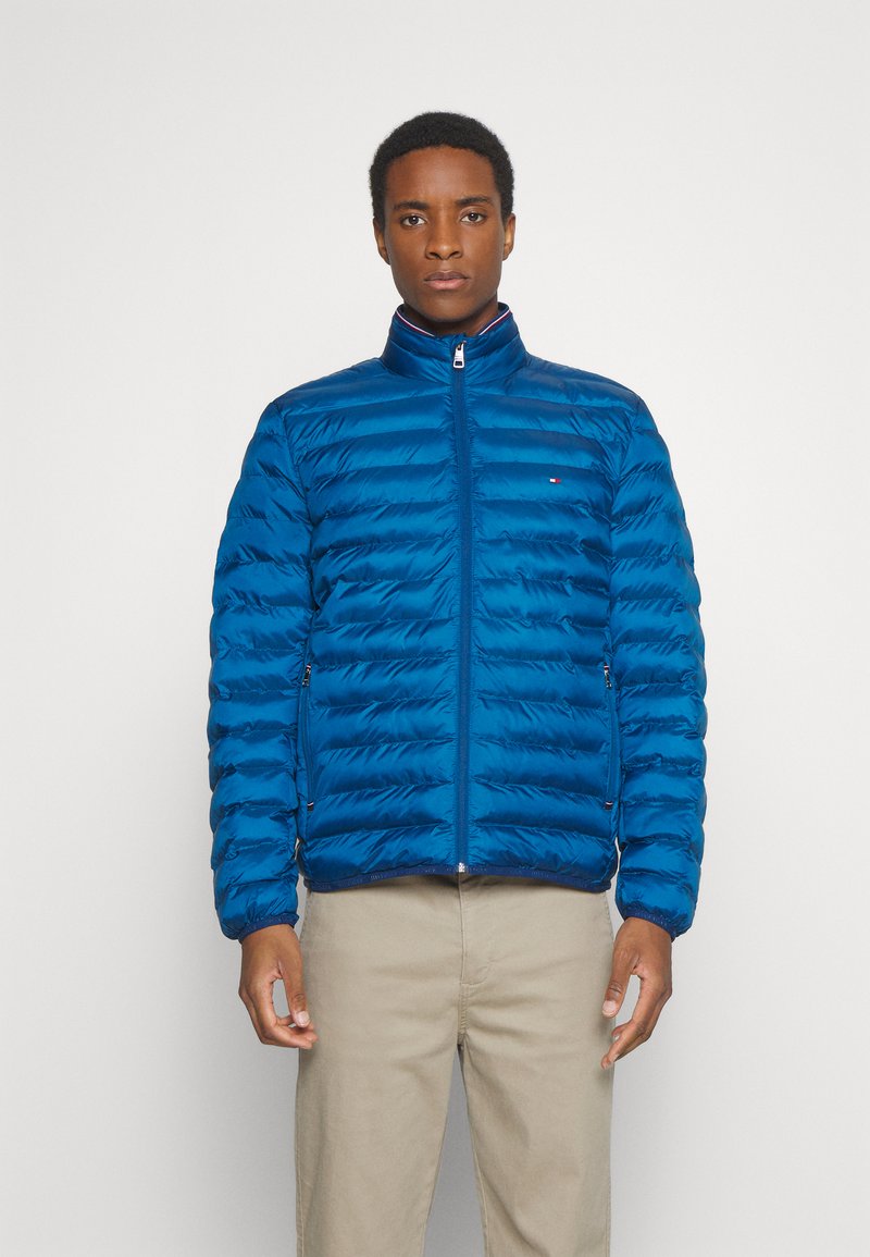 Blauwe pufferjack met hoge kraag, ritssluiting en horizontale stiksels. Voorzien van zijzakken en een logo op de borst. Lichtgewicht materiaal.