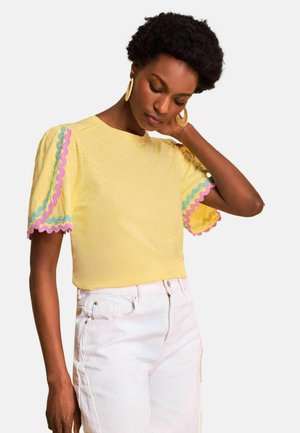 Woman with short curly hair wearing yellow short-sleeve top with pink and blue wavy trim and white high-waisted pants, looking down with hand on neck.