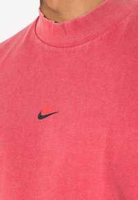 Krátký rukáv, vysoký límec tričko v jasně růžové bavlně s texturovaným povrchem. Na hrudi se nachází malé černé logo Nike.
