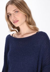 Granatowy sweter dzianinowy o luźnym kroju, szerokim dekolcie i fakturowanym wzorze. Posiada lekkie drapowanie od ramion.