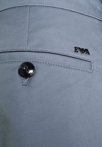 Svetlemodrá látková zadní kapsa s černým knoflíkem a malým černým logem. Hladká textura s jemnými detaily šití.