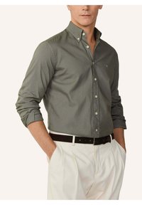 Mężczyzna w oliwkowo-zielonej koszuli na guziki z podwiniętymi rękawami, włożonej do białych plisowanych spodni z czarnym paskiem, ręce w kieszeniach.