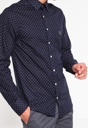Mężczyzna ubrany w granatową koszulę na guziki z długim rękawem, ozdobioną małym geometrycznym wzorem, oraz szare spodnie, z jedną ręką w kieszeni.