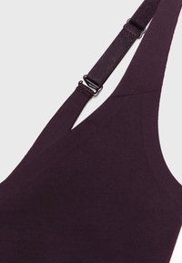 Gros plan sur une sangle ajustable en tissu violet foncé et une partie d’un vêtement, montrant la boucle en métal et le détail de la couture.
