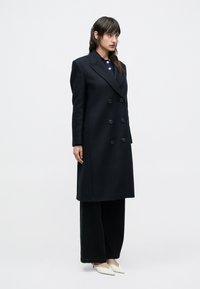 Vrouw staand in een lange zwarte dubbelgeknoopte jas, brede zwarte broek en witte pumps met spitse neus tegen een effen witte achtergrond.