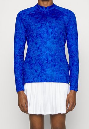 Persoon draagt een blauw patroon top met lange mouwen en een hoge kraag en een witte geplooide rok, staande tegen een effen achtergrond.