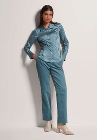 Satin-blaugrüne Bluse mit klassischem Kragen und durchgehender Knopfleiste, kombiniert mit strukturierten hellblaugrünen Cordhosen und beigen High Heels.