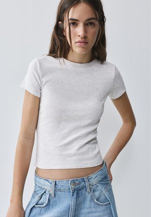 Junge Frau mit langen braunen Haaren, die ein schlichtes hellgraues kurzärmeliges T-Shirt und blaue Jeans trägt und mit einer Hand an der Hüfte steht.