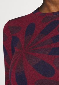 Detailní záběr na osobu, která má na sobě červený a tmavě modrý pletený svetr s abstraktními, vyzařujícími tvary na látce.