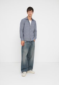 Svetlo modrá košeľa na gombíky z textúrovanej látky, skombinovaná s voľnými modrými džínami a bielymi teniskami, vytvárajúca ležérny outfit.