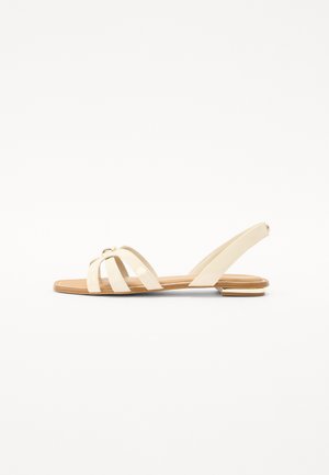 Beige flat sandals with crisscross straps, a pointed toe, and a slim ankle strap. Glossy finish with a gold-tone accent detail.