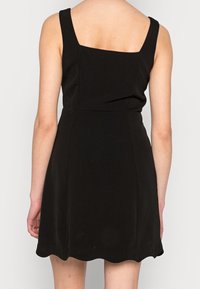 Black sleeveless dress with a square back, fitted bodice, and flared skirt. Smooth fabric and scalloped hem detail enhance the design.