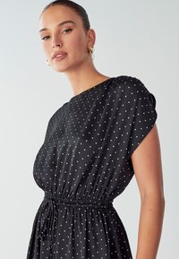 Schwarzes Kleid mit Punkten, kurzen Ärmeln, taillierter Taille und glattem Stoff. Verfügt über gleichmäßig verteilte weiße Punkte auf dem Kleidungsstück.