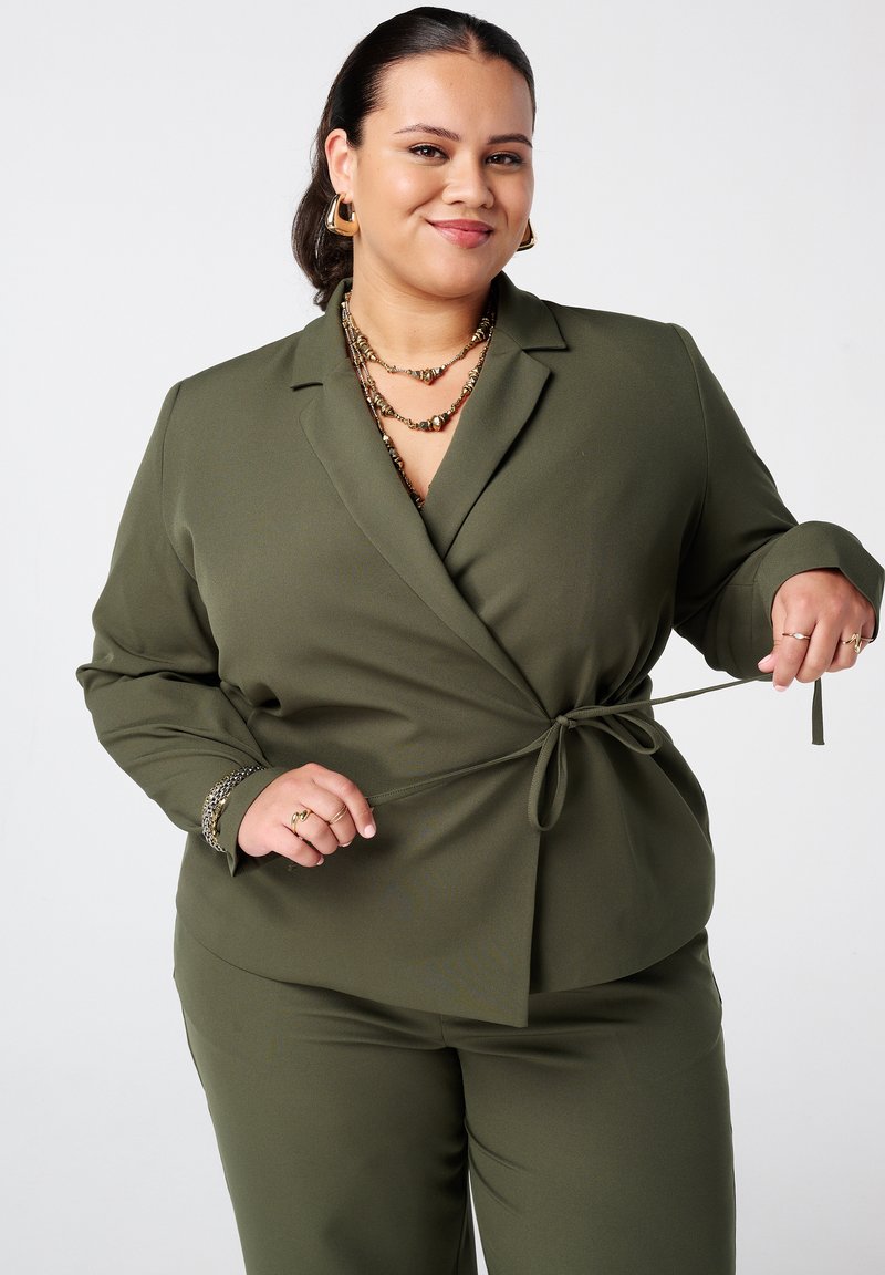 Olijfgroene twee-delige suit met een wikkelblazer met een strik taille en lange mouwen, gecombineerd met bijpassende maatbroeken. Textuurstof.