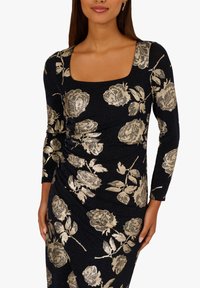 Donna che indossa un vestito nero aderente con motivo floreale metallico dorato e scollatura quadrata, mostrando il busto superiore e le braccia.