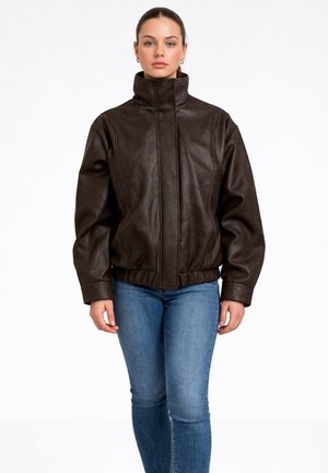 Woman wearing a dark brown leather jacket with high collar and blue jeans, standing against a plain white background.