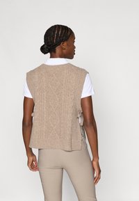 Vestă tricotată bej cu un model texturat în formă de cablu, părți laterale deschise și tiv cu striații, purtată peste o cămașă albă cu guler și pantaloni bej.