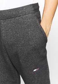 Dunkelgraue Jogginghose aus weichem Material, mit Seitentaschen und einem dezenten Logo in rot, schwarz und blau near der linken Tasche.