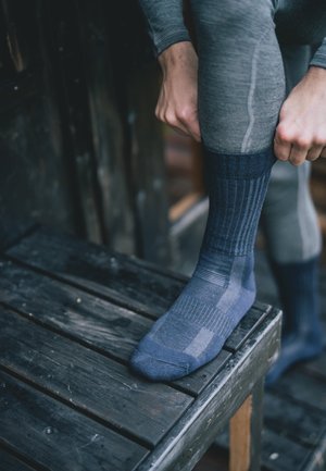 Chaussettes bleu foncé en tissu texturé avec un motif, dotées de bords côtelés et de orteils renforcés, portées en se tenant debout sur un tabouret en bois sombre.
