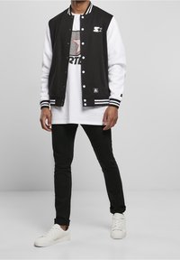 Veste universitaire noir et blanc avec poignets côtelés rayés, t-shirt noir avec imprimé graphique, jean skinny noir et baskets blanches.