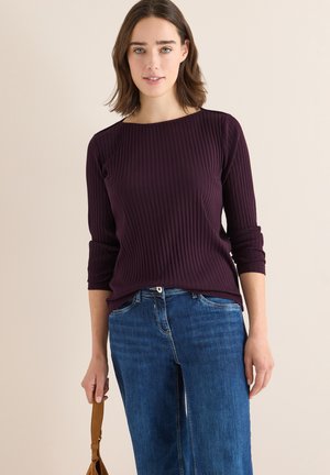 Top viola a coste con maniche tre quarti, abbinato a jeans blu a gamba larga. La modella tiene una borsa beige, su uno sfondo neutro.