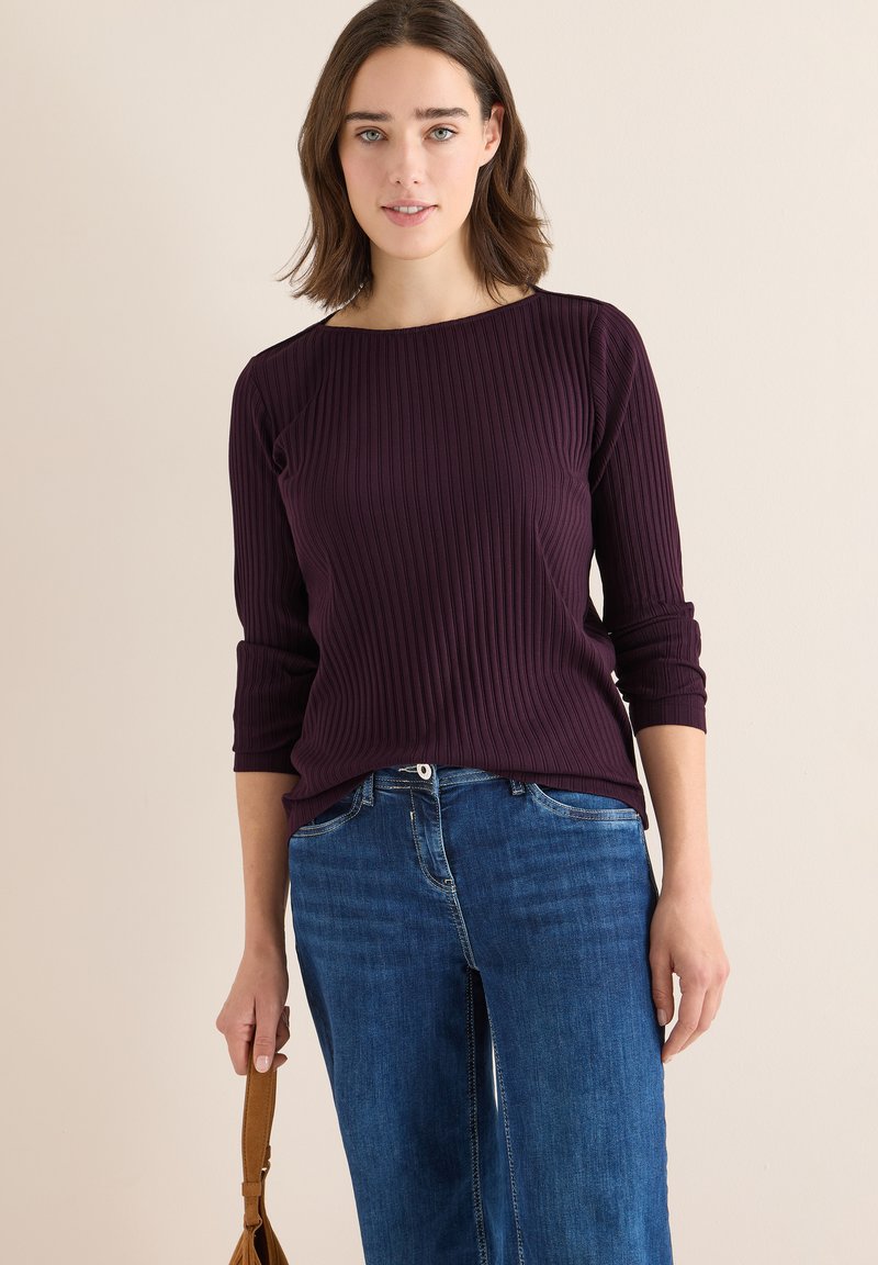 Ribbade lila toppar med trekvartslånga ärmar, ihop med blå vida jeans. Modellen håller en beige väska, mot en neutral bakgrund.