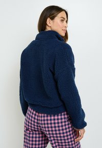 Donna con capelli castani che indossa una giacca in pile blu navy e pantaloni a quadri rosa e blu navy, rivolta di lato su uno sfondo neutro.