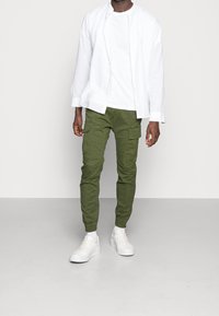 Weißes Langarmhemd über einem schlichten weißen T-Shirt, grüne Cargo-Hosen mit Seitentaschen und weiße Sneaker auf einem grauen Boden.