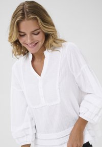 Blusa bianca in cotone con un motivo testurizzato, caratterizzata da una placca con bottoni, maniche a sbuffo di lunghezza intermedia e finiture decorative lungo l'orlo.