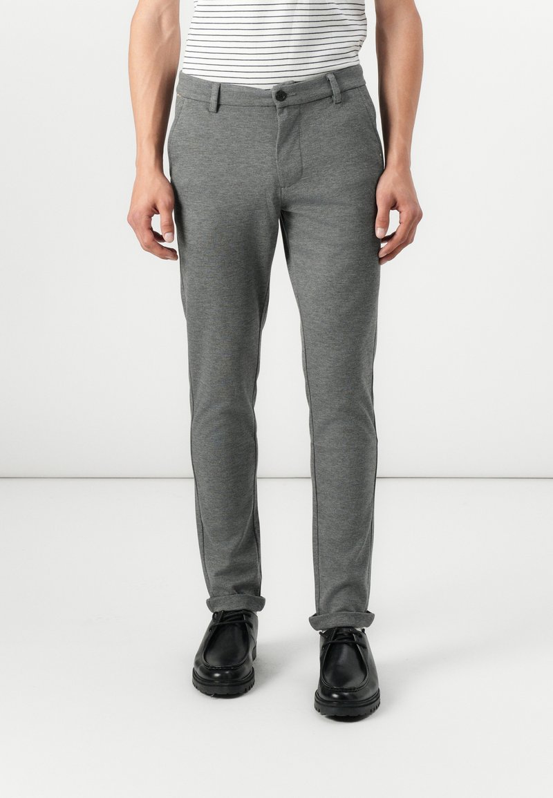 Graue, maßgeschneiderte Hose aus weichem Stoff, mit flacher Front, seitlichen Taschen und schlankem Schnitt. Getragen mit schwarzen Schuhen.