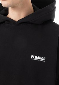 Hanorac negru cu glugă, fabricat dintr-un material moale, având un logo alb "PEGADOR" și textul "CERTIFICAT AUTENTIC STREETWEAR" pe piept.