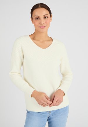 Jersey de manga larga con escote en V en color crema, con tejido texturizado en la parte superior y puños acanalados. Material suave, corte relajado, combinado con unos vaqueros azul claro.