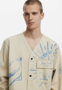 Cămașă oversized bej cu imprimeuri grafice albastre, inclusiv un design cu soare, și un buzunar frontal. Prezintă un decolteu adânc în V și închidere cu nasturi.