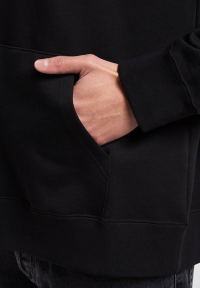 Black hoodie featuring a spacious front pocket, ribbed cuffs, and a textured fabric. Detail of a hand resting in the pocket shown.