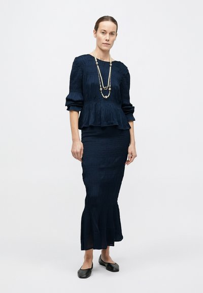 Femeie în picioare, purtând o bluză texturată albastru închis și o fustă lungă asortată, balerini negri și un colier lung cu mărgele, pe un fundal simplu.