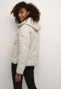 Quiltet lys beige jakke med hætte, der har et diamantmønster og et enkelt logo-patch på venstre ærme. Båret med mørke jeans.