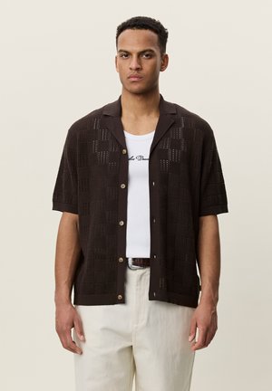 Jeune homme portant une chemise marron foncé à manches courtes boutonnée sur un débardeur blanc et un pantalon beige clair sur un fond uni.