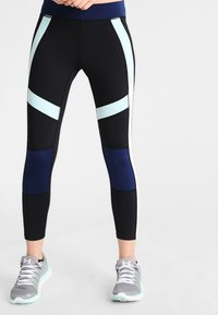 Kvinna iklädd svarta leggings med ljusblå och mörkblå färgblockeringsmönster samt grå sportskor, stående med händerna vid midjan.