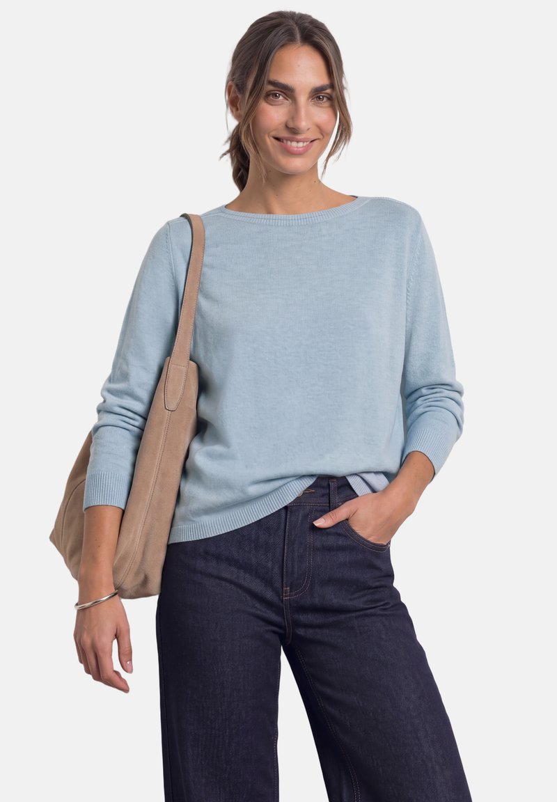 Femme aux cheveux attachés porte un pull bleu clair et un jean foncé, porte un sac à épaule beige, sourit avec une main dans la poche.