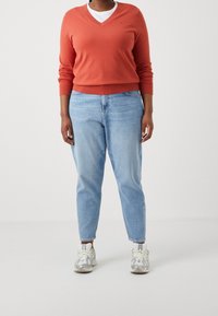 Pull col en V corail avec poignets côtelés, porté sur un t-shirt blanc, associé à un jean bleu clair droit et des baskets grises.