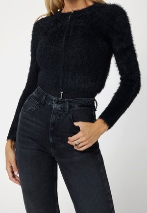 Femme portant un pull à fermeture éclair noir et pelucheux associé à un jean noir taille haute, une main dans la poche.