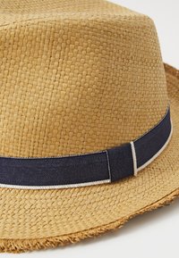Sombrero de paja trenzada en color beige, con una banda de tela azul marino y borde blanco. Ala ancha con borde deshilachado. Diseño texturizado y tejido.