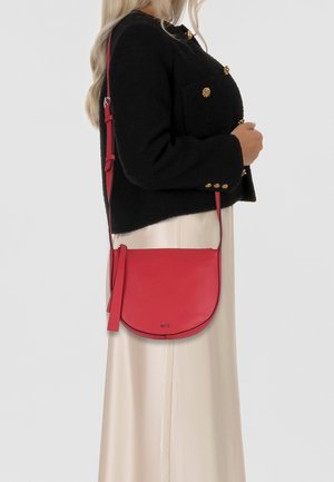 Red leather crossbody bag with pebbled texture, curved shape, and adjustable strap, featuring a subtle logo in the front.