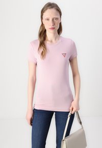 T-shirt rosa a maniche corte con design aderente e un piccolo logo a triangolo rosso sul petto, abbinato a jeans blu scuro e una borsa beige.