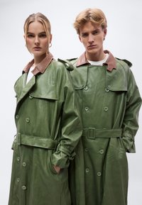 Πράσινα δερμάτινα παλτό τύπου τρεντς με καφέ λεπτομέρειες στον γιακά, διπλό μπροστινό κουμπί, μέση με ζώνη και πλαϊνές τσέπες, φορεμένα από δύο μοντέλα.