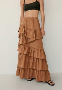 Jupe maxi brun à plusieurs niveaux avec des couches volantées, taille élastique et texture de tissu douce, associée à un haut court sombre et des sandales.