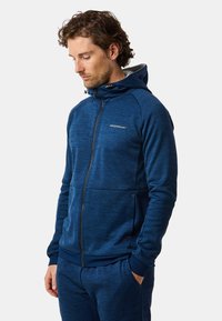 Mies, jolla sininen vetoketjullinen huppari ja siihen sopivat housut, kädet taskuissa, katsoo alas, seisoo yksiväristä vaaleaa taustaa vasten.