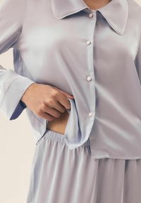 Jasnoniebieski zestaw satynowych piżam, składający się z koszuli z kołnierzykiem na guziki z kieszonką oraz spodni z elastycznym pasem, prezentujący gładką teksturę i delikatny blask.